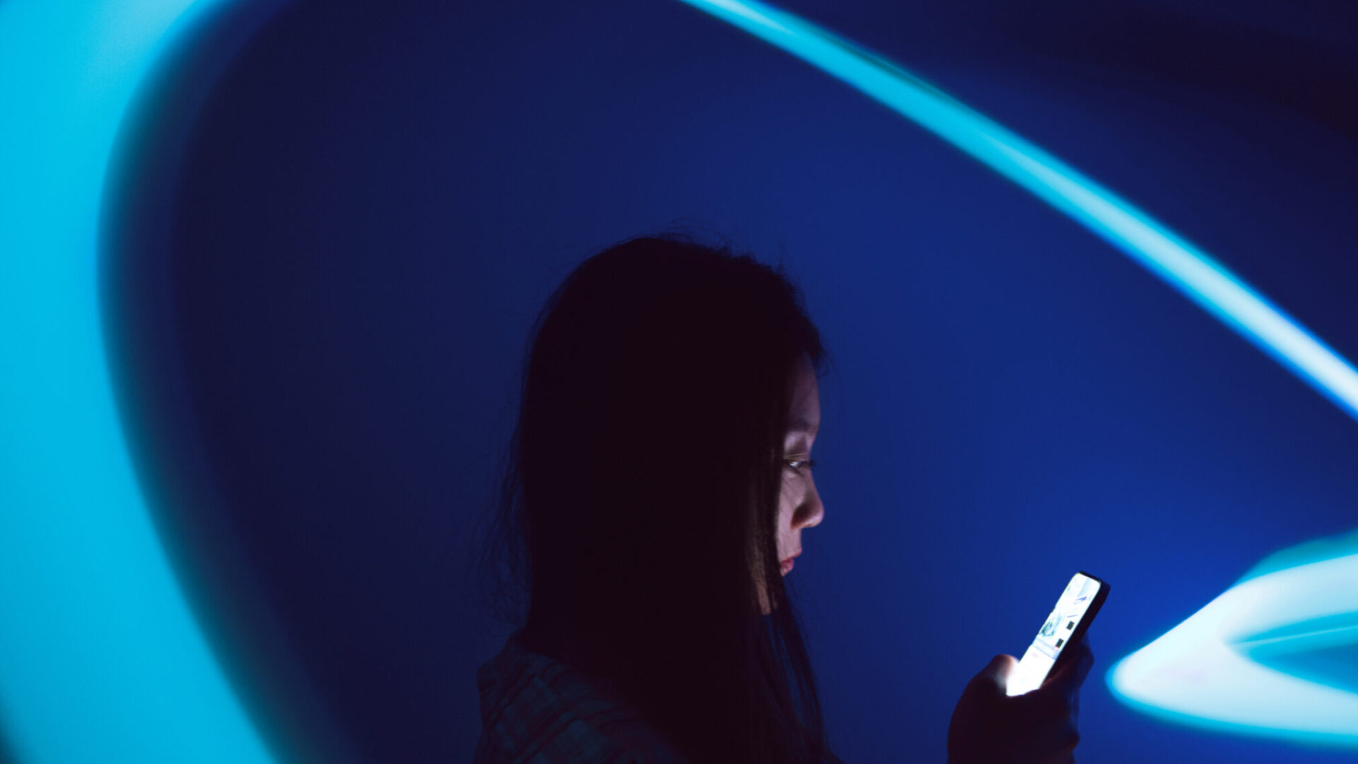 A person looking at a smartphone against a dark blue background with sweeping turquoise light trails.