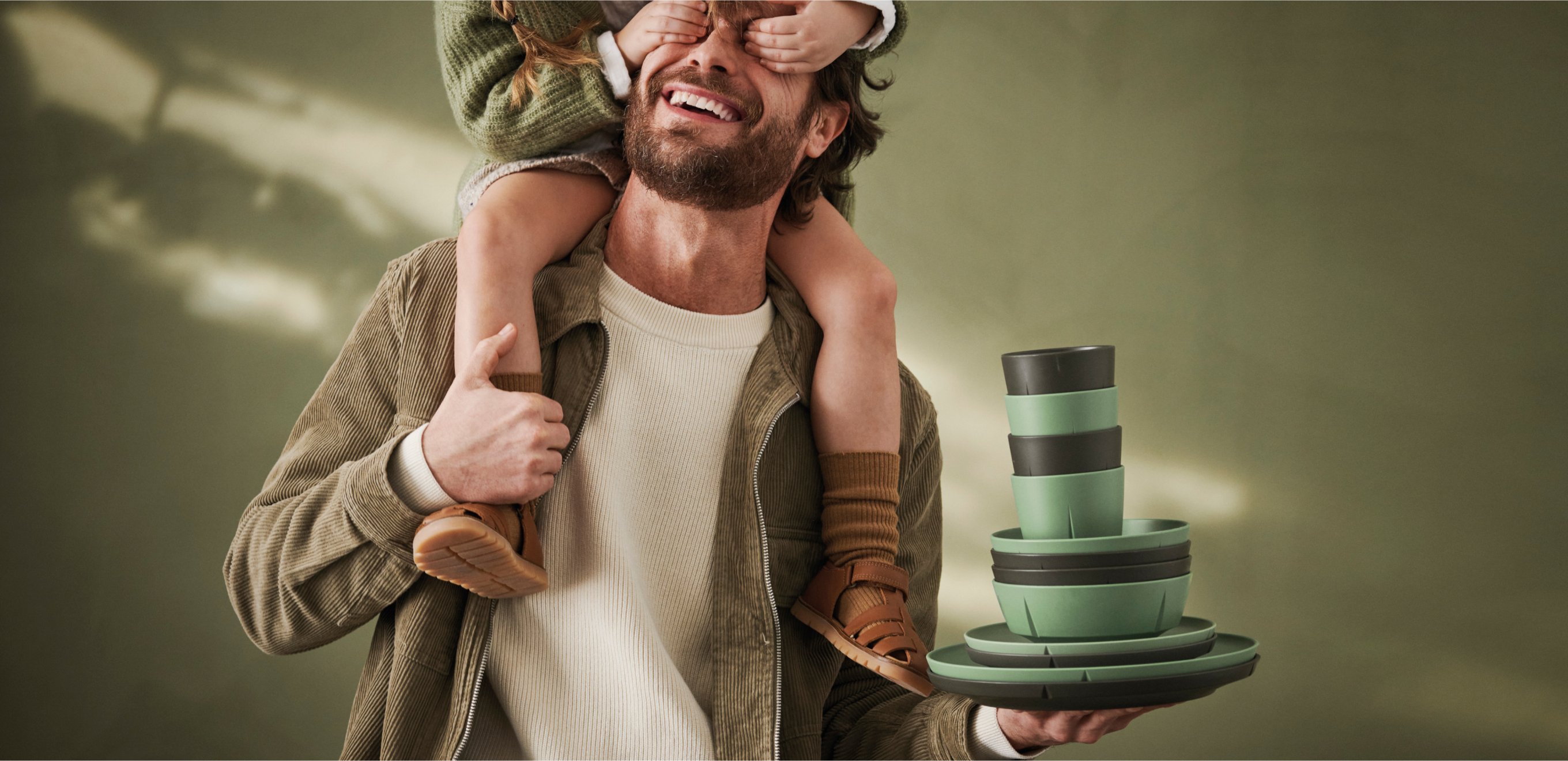 A smiling man carrying a child on his shoulders and balancing stacked green and black tableware.