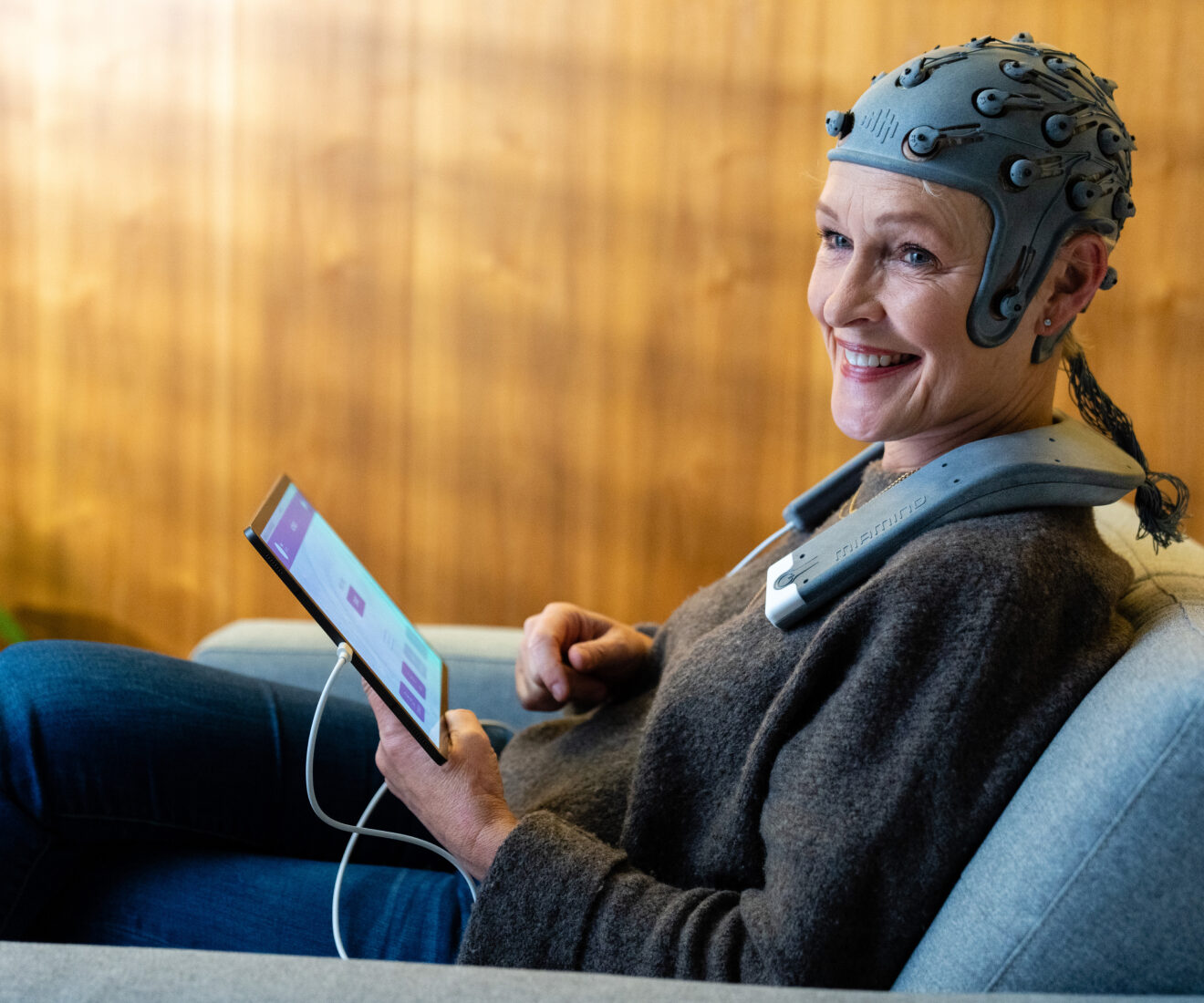 Smiling woman wearing a sensor cap and holding a tablet while seated on a sofa.