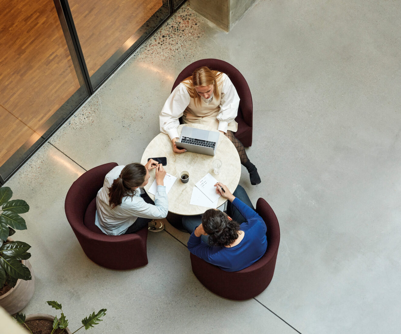 Overhead view of three colleagues working around a small round table with a laptop, notes and coffee.