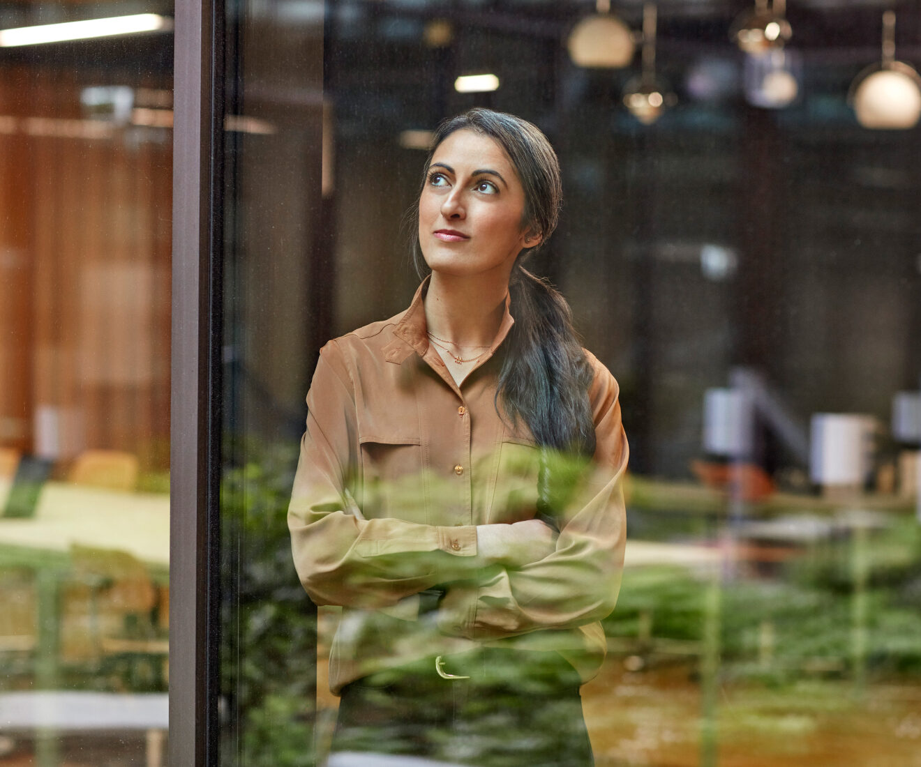 Woman in a brown shirt standing with arms crossed by an office window, looking upward.