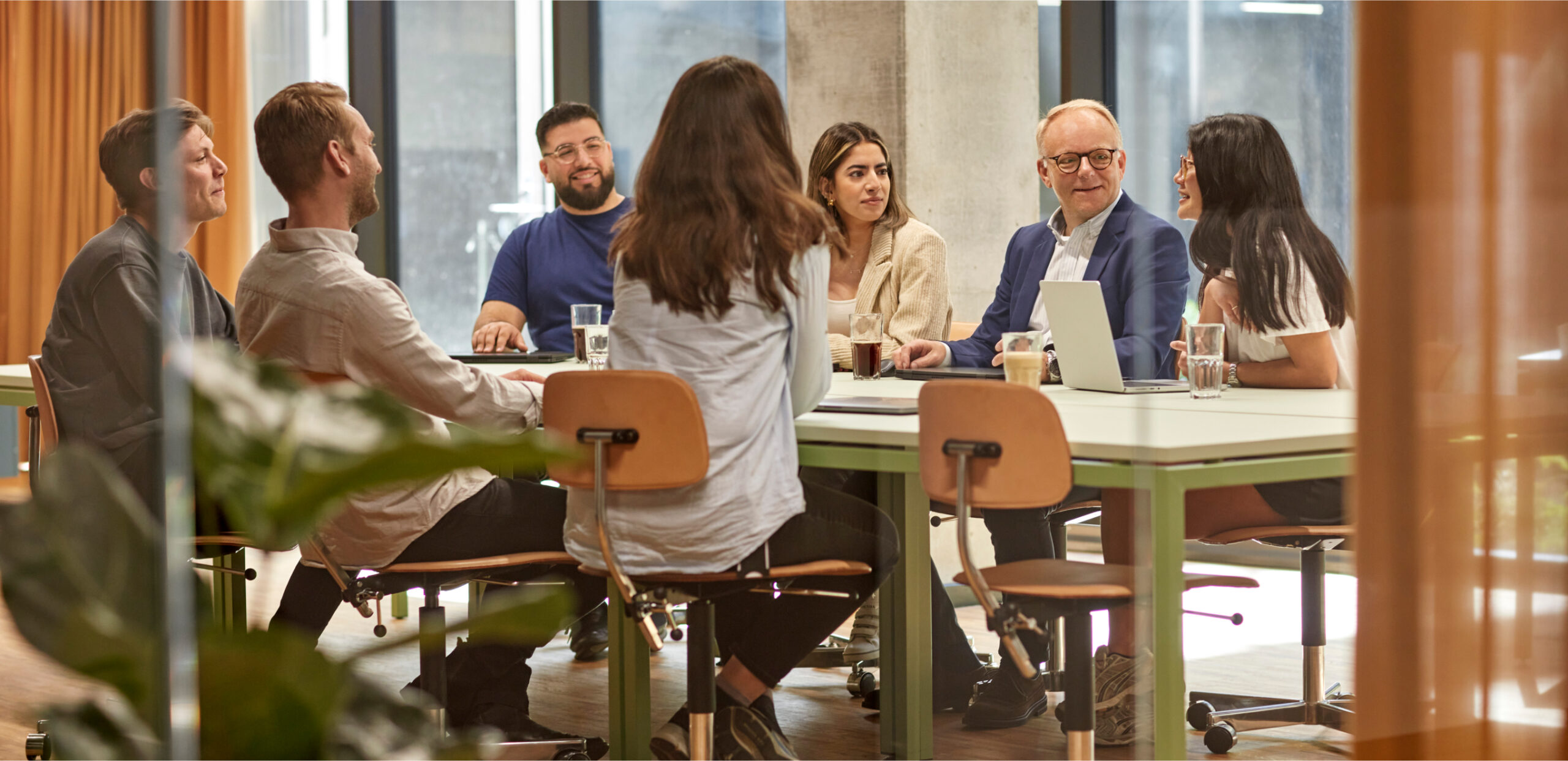 Seven people talking around a meeting table with a laptop and drinks in a bright office.