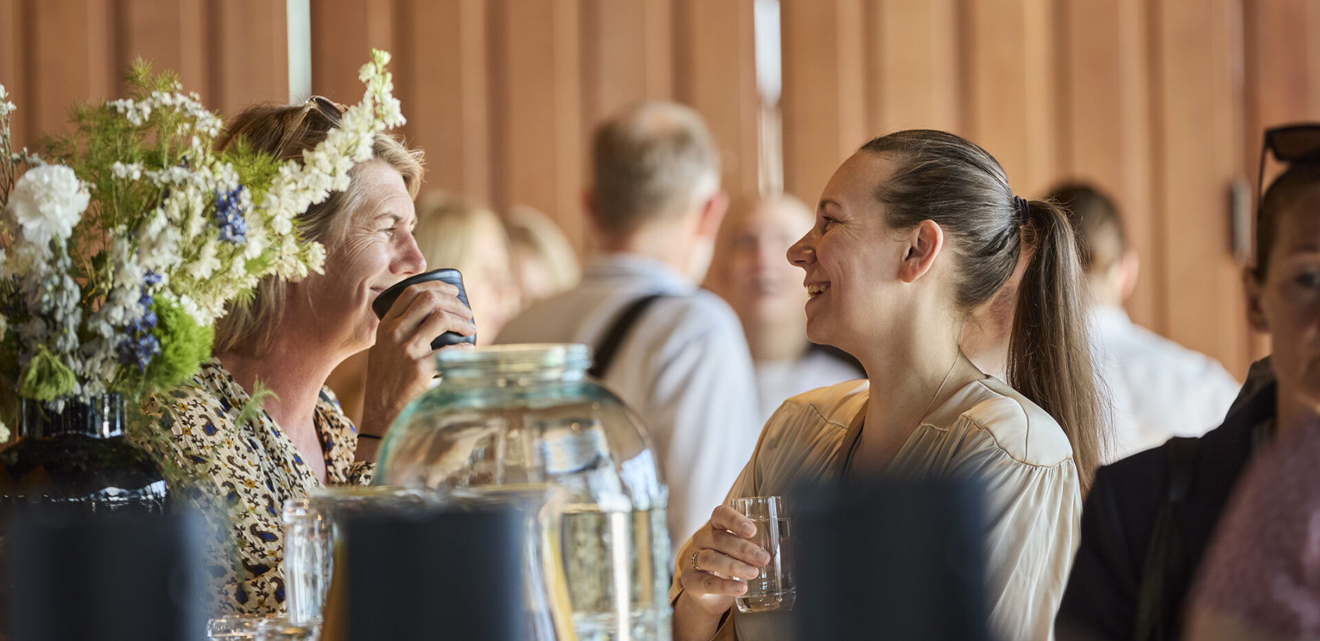 Two women smiling and chatting over drinks at an indoor gathering, with flowers and glassware in the foreground.