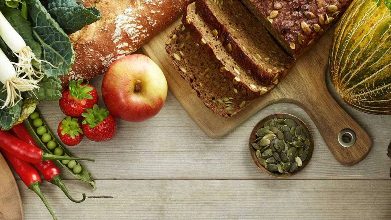 Bread, fruit, vegetables and pumpkin seeds arranged on a wooden table.