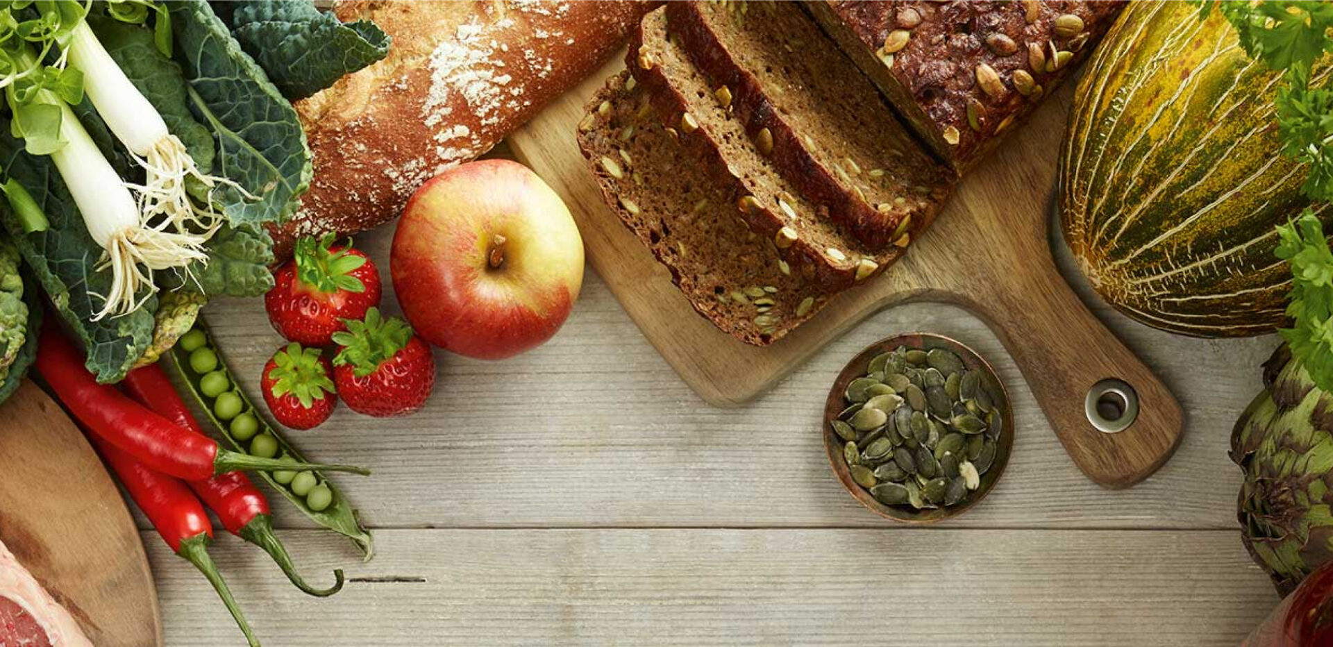 Bread, fruit, vegetables and pumpkin seeds arranged on a wooden table.