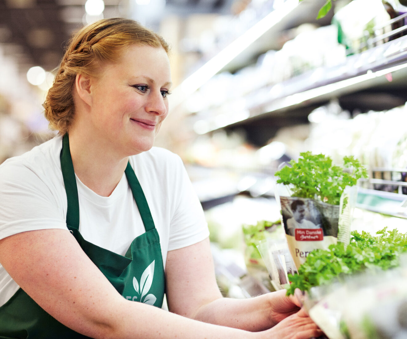 A woman in a green apron arranging potted herbs on a supermarket shelf.