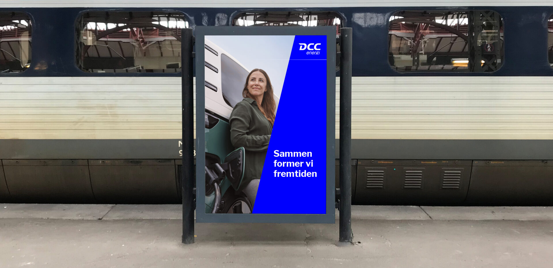 DCC Energi advertisement on a train platform showing a woman beside a truck and the slogan “Together we shape the future”.