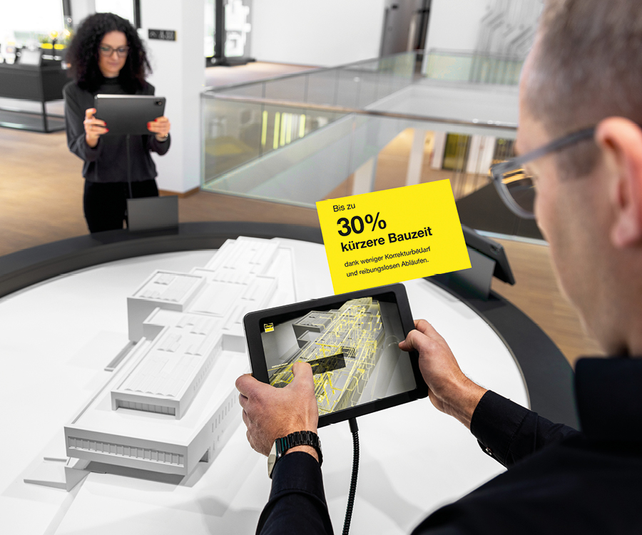 Two people using tablets to explore a digital architectural model in a modern exhibition space, with a yellow German sign highlighting up to 30% shorter construction times.